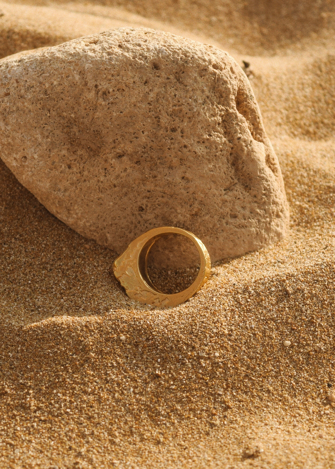 Side view of the cracked gold ring by Divon Jewelry, 18K gold-plated, resting on sand near a natural stone for an earthy presentation.